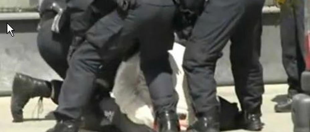 Das Video zeigt mehrere Beamte, die einen Mann in Handschellen am Boden halten. Der Beamte am linken Bildrand tritt darin mehrfach zu.
