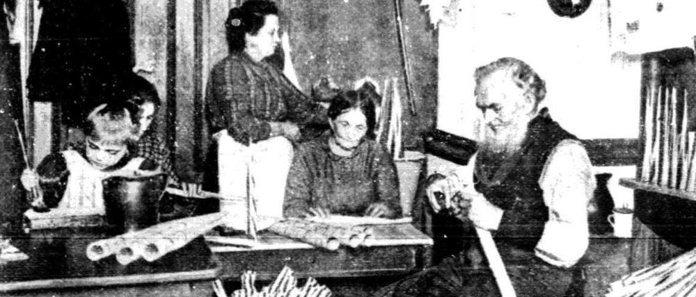 Eine fünfköpfige Familie sitzt am Tisch ihrer Einzimmer-Wohnung und fertigt Papptröten für Jahrmärkte. Auch ein Kind hilft bei der Heimarbeit mit.