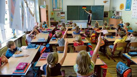 Blick ins Klassenzimmer einer Grundschule, Schüler melden sich,  vorn an der Tafel steht die Lehrerin.
