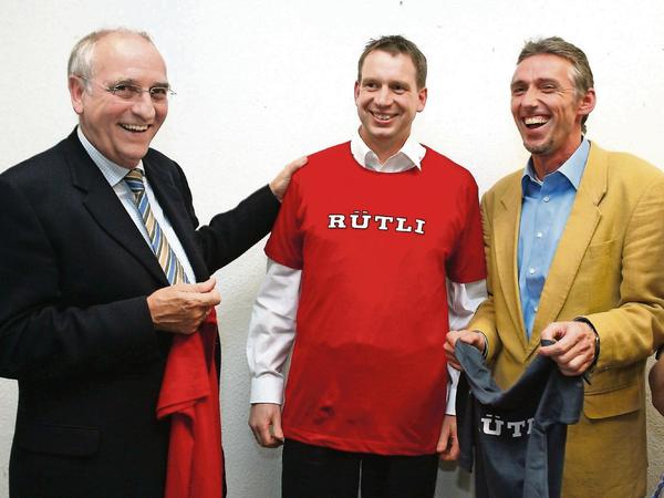 Im Herbst 2006 streifen Bildungssenator Klaus Böger (links) und der Rütli-Interimsschulleiter Helmut Hochschild dem neuen Leiter Aleksander Dzembritzki ein Shirt aus der Rütli-Wear-Kollektion über. 