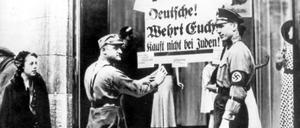 Deutschland 1938 – ein Nazisoldat klebt Plakate ans Schaufenster eines jüdischen Geschäfts. Zuletzt forderten Antisemiten mehrfach den Boykott jüdischer Unternehmen.