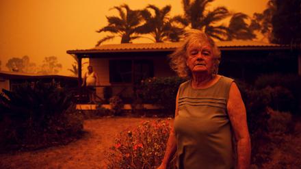 Eine Frau steht vor ihrem Haus in Australien, während Wind Rauch und Asche herbei trägt. 