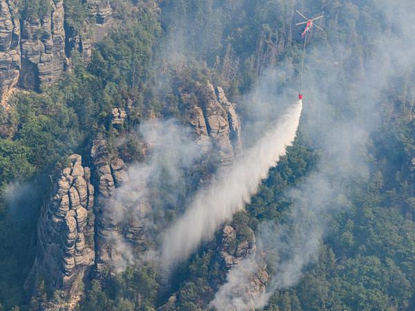 Ein Lastenhubschrauber aus Österreich fliegt mit einem Löschwasser-Außenlastbehälter um einen Waldbrand im Nationalpark Sächsische Schweiz zu löschen.