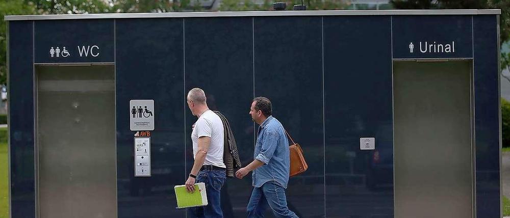 Zwei Männer gehen in Köln an einer öffentlichen Toilette vorbei. Frauen müssen für die Benutzung dieser Toilettenanlagen 50 Cent zahlen, während Männer gratis das Urinal benutzen dürfen.