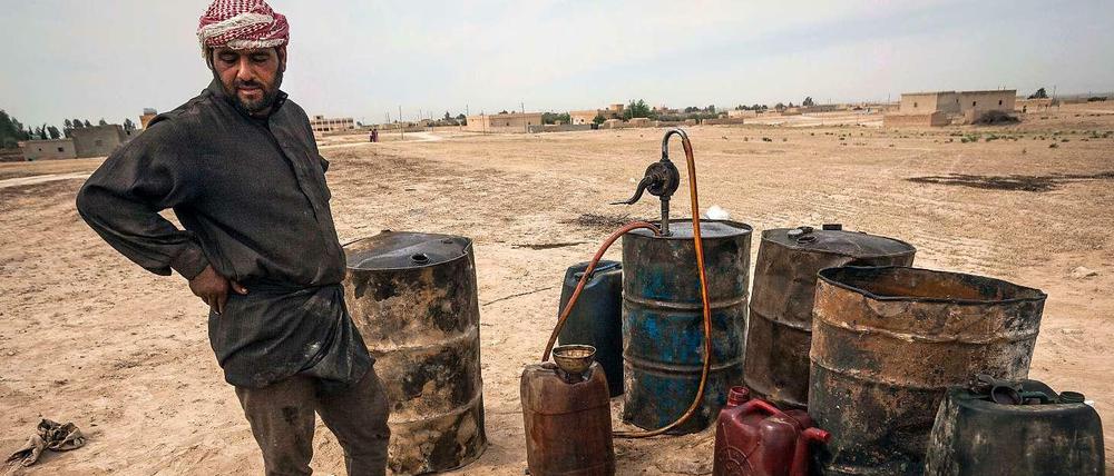 Das Bild zeigt einen Mann in einfacher Kleidung mit einem roten Palästinenserschal um den Kopf. Er steht neben verbeulten und verrosteten Fässern und Kanistern im Nichts einer sandigen Ebene.
