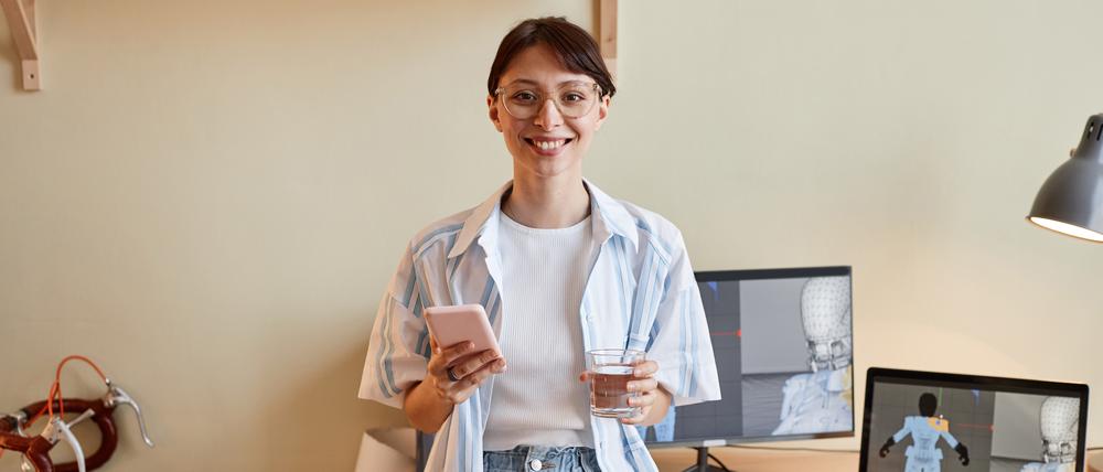 Eine lächelnde junge Frau mit Smartphone und Laptop am digitalen Arbeitsplatz.