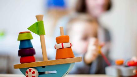 Auf einem Tisch in einem Kindergarten steht ein Holzspielzeug auf einem Tisch, im Hintergrund ist ein Kleinkind zu sehen.