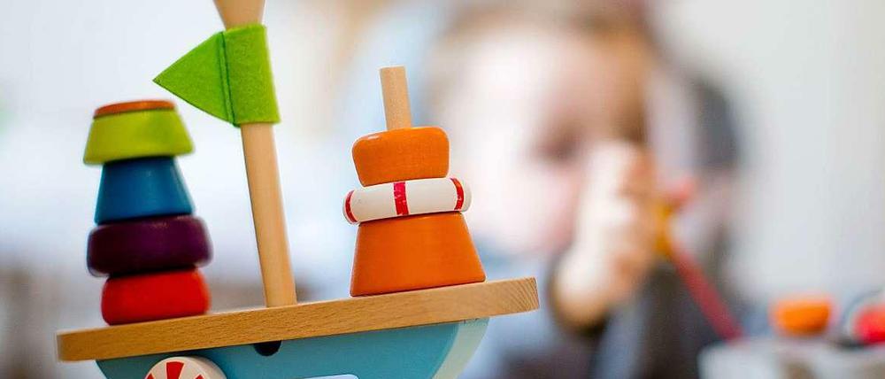 Auf einem Tisch in einem Kindergarten steht ein Holzspielzeug auf einem Tisch, im Hintergrund ist ein Kleinkind zu sehen.