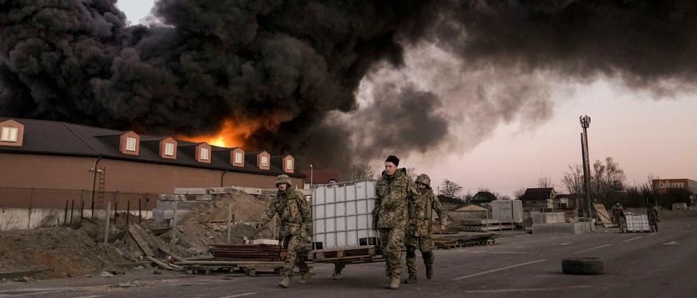 Ukrainische Soldaten in der Nähe von Kiew tragen einen Container, hinter ihnen steht ein Lagerhaus nach einem Bombenanschlag in Flammen. 