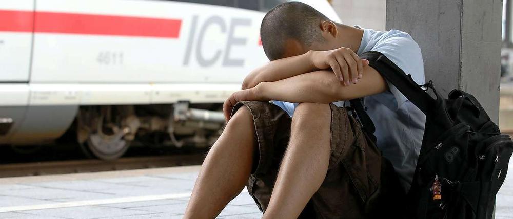 Ein junger Mann in kurzer Hose sitzt auf einem Bahnsteig, den Kopf auf die Arme gestützt. Im Hintergrund steht ein ICE-Zug.