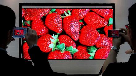 Auf einem rieseigen Bildschirm sind strahlend rote Erdbeeren zu sehen. Davor zwei Betrachter, die den Fernsehbildschirm fotografieren.