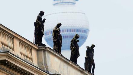 Auf dem Dachsims eines Gebäudes stehen vier Skulpturen, im Hintergrund die Aussichtskugel eines Fernsehturms.