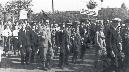 Männer und Frauen marschieren über einen Platz, dabei halten sie ein Schild mit der Aufschrift Walther de Gruyter hoch.