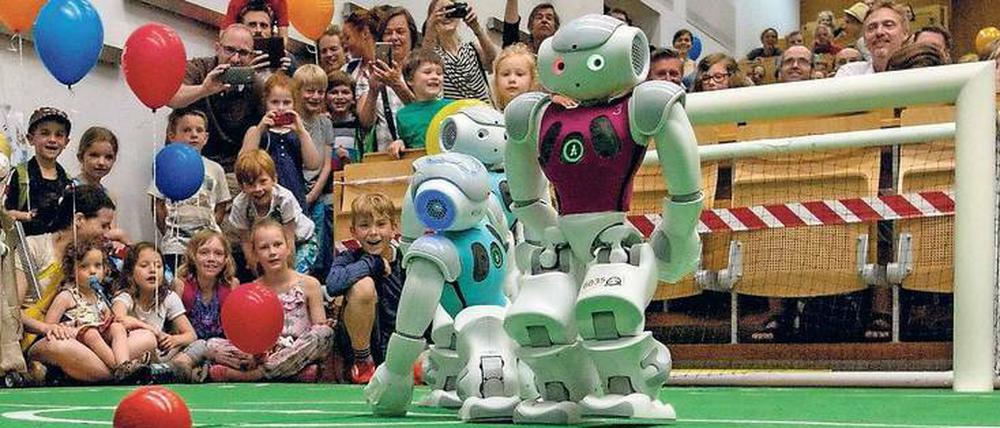 Roboter spielen Fußball, Kinder und Erwachsene schauen ihnen dabei zu.