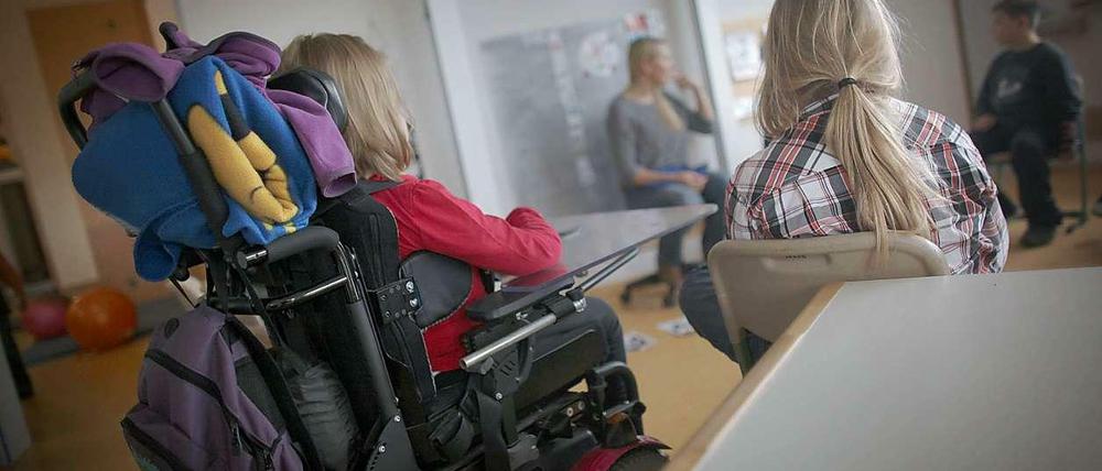 In einem Klassenraum sitzt eine Schülerin im Rollstuhl neben einer Schülerin, die auf einem Stuhl sitzt.