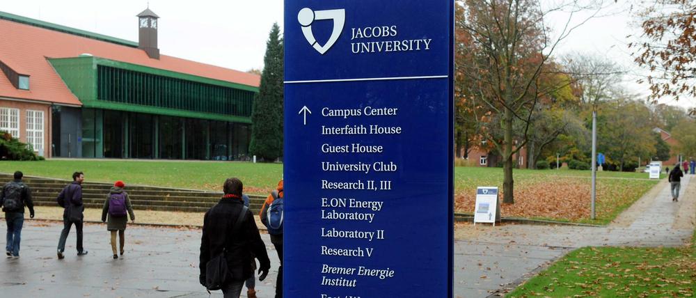 Studierende laufen bei herbstlichem Regenwetter über einen Campus, im Vordergrund ein Hinweisschild zu Unigebäuden.