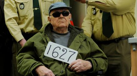 John Demjanjuk sitzt mit einer Nummer in der Hand in einem Rollstuhl, hinter ihm stehen Justizangestellte.