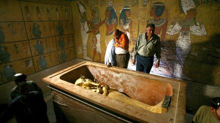 Drei Männer stehen in einem mit Wandbildern geschmückten Raum neben einem Sarkophag.