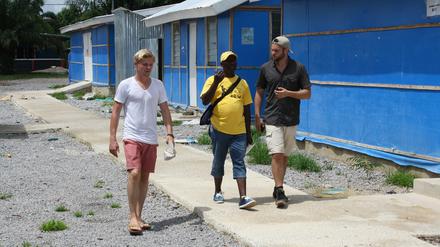 Drei Männer gehen einen Weg entlang, im Hintergrund sind Gebäude in Schnellbauweise zu sehen.