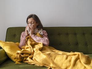 Eine Frau sitzt mit Decke auf dem Sofa und schnäuzt sich die Nase.