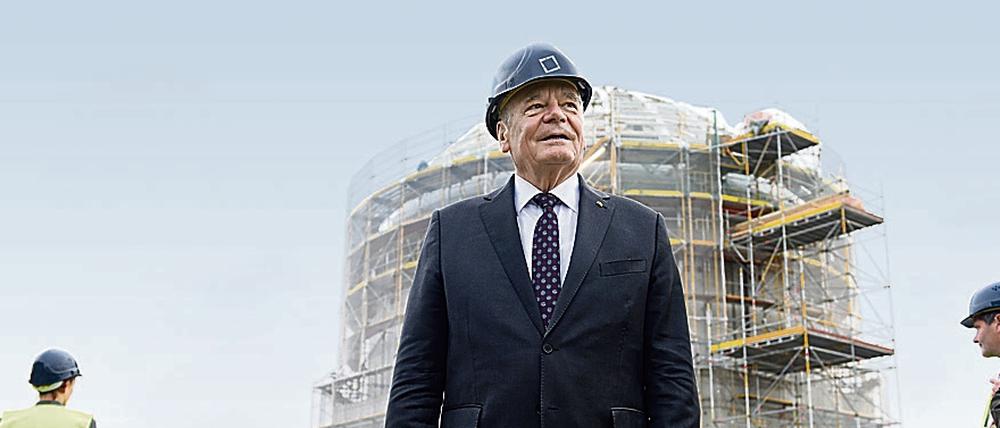 Bundespräsident Joachim Gauck steht mit herkömmlichen Arbeitsschuhen und Helm bekleidet auf dem Dach des Gebäudes vor der Kuppel. 