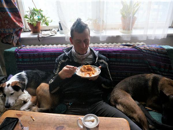 Burkhard Garweg auf einem neueren Foto – auf der Couch mit zwei Hunden und einem Teller Spirelli mit Tomatensoße in der Hand.