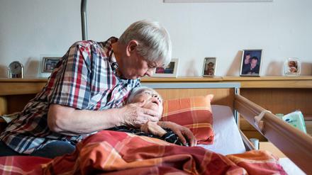 Ein pflegender Ehemann nimmt in seiner Wohnung seine an Demenz und Parkinson erkrankte Ehefrau in den Arm.