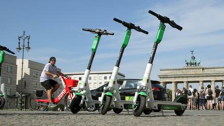 E-Tretroller der US-amerikanischen Firma Lime stehen vor dem Brandenburger Tor, während im Hintergrund ein Mann auf einem E-Bike des Anbieters Uber vorbeifährt. (Archivbild)