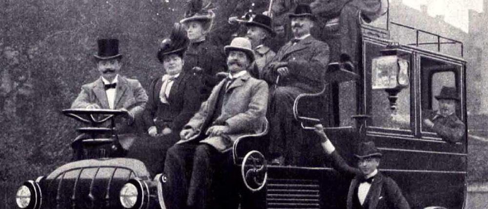 Auf einem kutschenähnlichen Fahrzeug sitzen Männer und Frauen auf dem Führerstand. Es handelt sich um einen Elektrobus mit Akkubetrieb, der 1899 von der Firma Kühlstein in Berlin gebaut wurde.