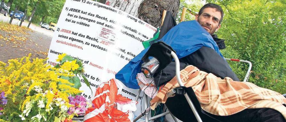 Unterm Baum. Firas Maraghy trinkt seit einem Monat nur Wasser. Er streikt für ein Reisedokument für seine Tochter. Unterstützer bringen ihm Blumen und Kerzen. 