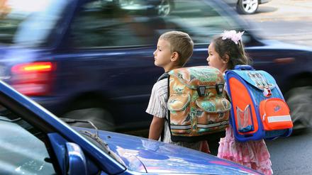 Schulkinder am Straßenrand, vor denen ein Auto vorbeirast, Thema Schulweg-Sicherheit.