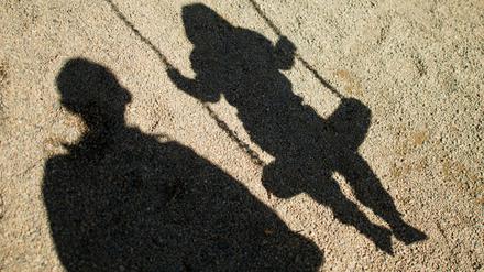Der Schatten von einem Mann und einem schaukelnden Kind fallen auf Sand auf einem Spielplatz. (Symbolbild)