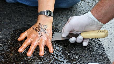 Ein Polizist löst die festgeklebte Hand eines Aktivisten der "Letzten Generation" in Berlin. Auf der Hand steht der Slogan "Öl sparen statt bohren".