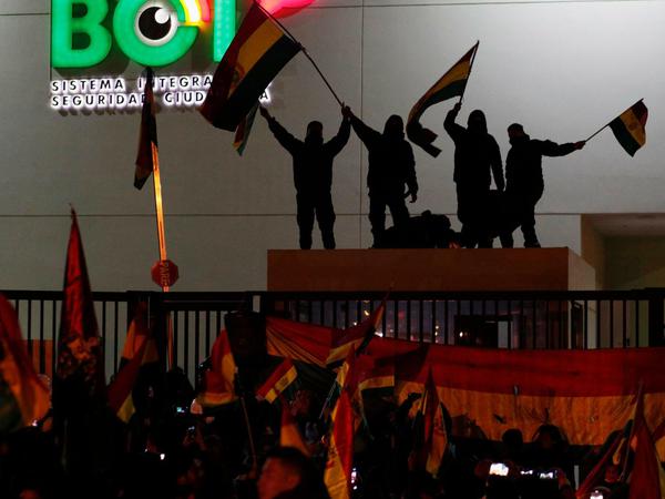 Polizisten, die auf dem Dach eines Sicherheitsstandes auf einer Polizeistation stehen, schwenken bolivianische Flaggen, im Vordergrund stehen Demonstranten. Evo Morales war auch durch den fehlenden Rückhalt der Sicherheitskräfte zum Rücktritt gezwungen worden. 