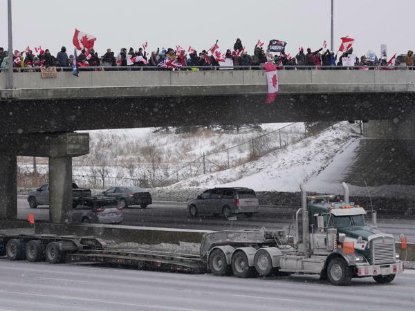 Unterstützer mit kanadischen Flaggen stehen auf einer Überführung und jubeln den vorbeifahrenden Lastwagen zu. 
