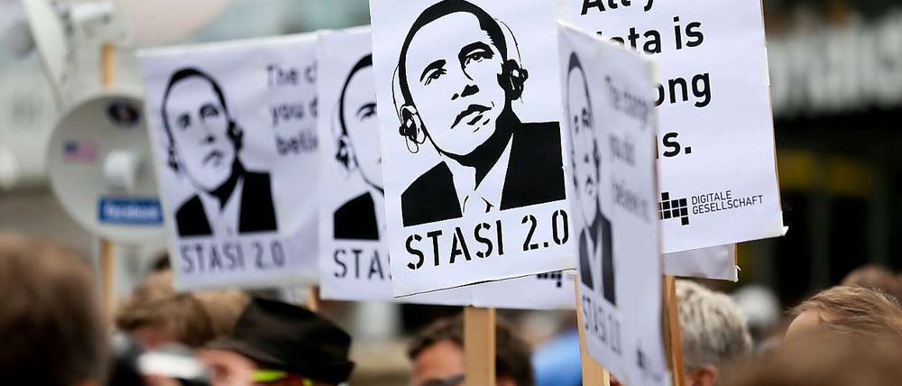 Demonstranten halten bei Protesten während des Besuchs von Barack Obama in Berlin am Checkpoint Charlie Plakate hoch, auf denen Obama mit Kopfhörern zu sehen ist.