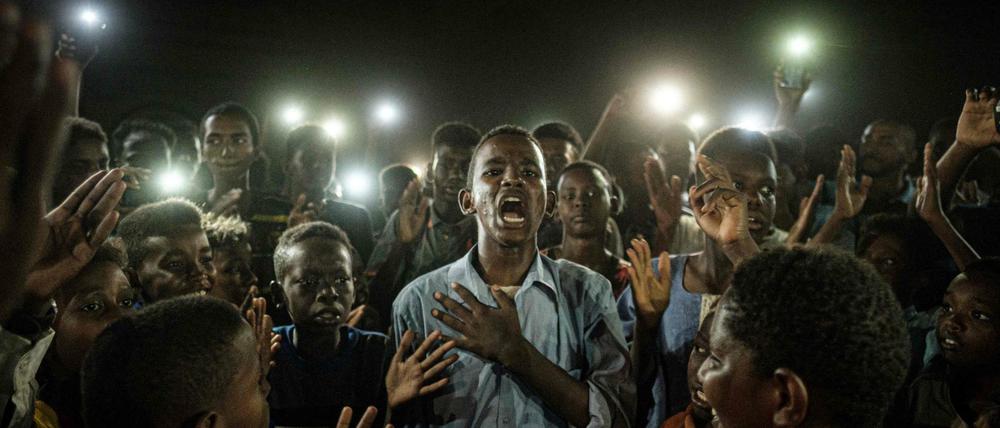 Das Bild zeigt, wie ein junger Sudaner umgeben von anderen Demonstranten im Dunklen inbrünstig ein Gedicht rezitiert - erhellt wird das Bild nur von Handylichtern der Umstehenden.