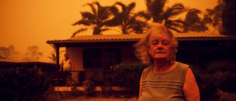 Eine Frau steht vor ihrem Haus in Australien, während Wind Rauch und Asche herbei trägt. 