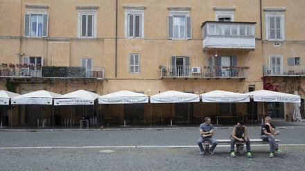 Drei Männer sitzen vor einem geschlossenen Restaurant auf der Piazza Navona in Rom auf einer Bank. 