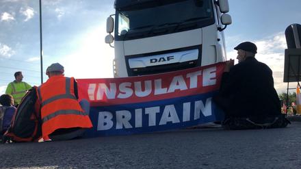 Zwei Männer sitzen vor einem Lastwagen und spannen einen Banner mit der Aufschrift "Insulate Britain".