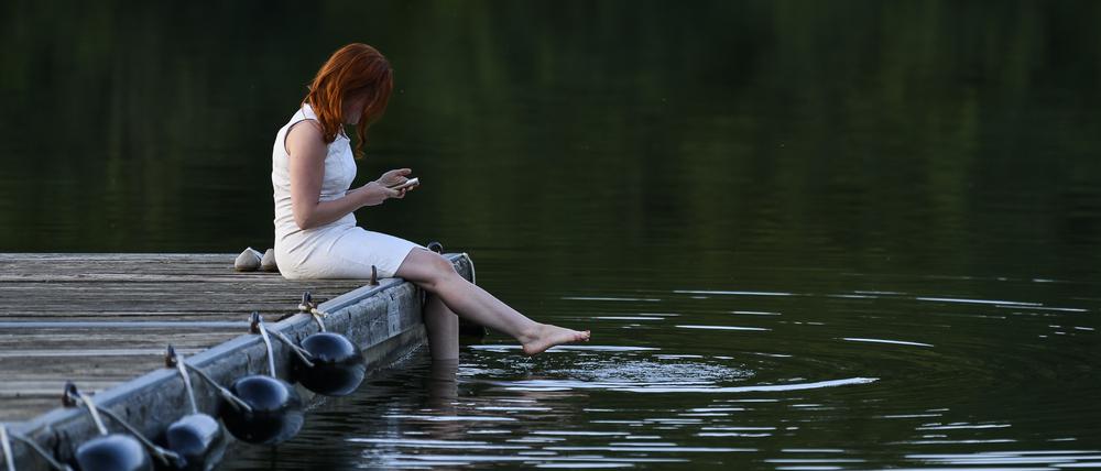 Eine Frau sitzt am Bucher Stausee im weißen Kleid auf einem Steg und schaut auf ihr Handy.