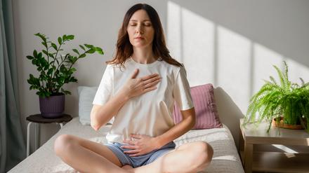 Pretty female sitting on bed in lotus posture with hands on her chest and belly doing pranayama techniques. Doing yoga at home. Self care and positive emotions.
