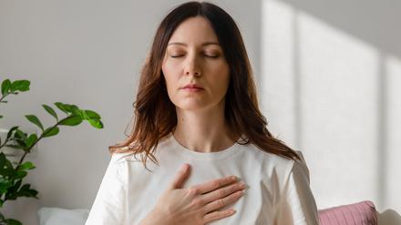 Pretty female sitting on bed in lotus posture with hands on her chest and belly doing pranayama techniques. Doing yoga at home. Self care and positive emotions.