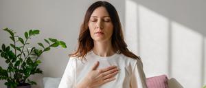 Pretty female sitting on bed in lotus posture with hands on her chest and belly doing pranayama techniques. Doing yoga at home. Self care and positive emotions.