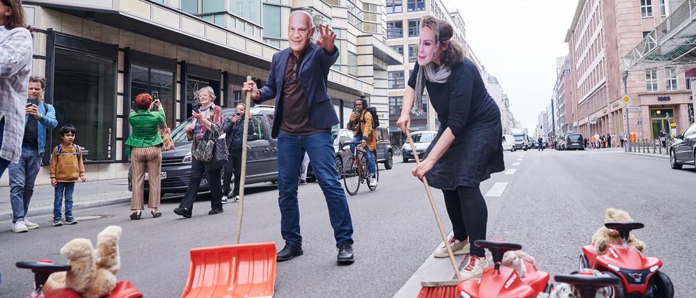 Ein Demonstrant mit einer Kai-Wegner-Maske und eine Demonstrantin mit Manja-Schreiner-Maske kehren symbolisch auf der Friedrichstraße die Bobbycars aus dem Weg.