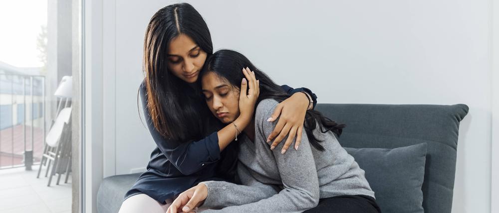 Eine junge Frau tröstet ihre Schwester auf dem Sofa im Wohnzimmer.
