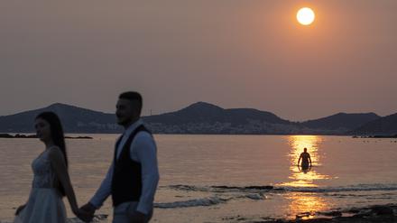 Ein frisch verheiratetes Paar posiert bei Sonnenuntergang für Fotos, während ein Mann ein Bad im Wasser in Lagonisi südöstlich von Athen nimmt.