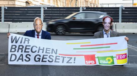 Klimaaktivisten sitzen auf einer Straße und haben sich als Kanzler Olaf Scholz (SPD) und Verkehrsminister Volker Wissing (FDP) verkleidet.