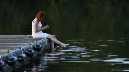 Eine Frau sitzt am Bucher Stausee im weißen Kleid auf einem Steg und schaut auf ihr Handy.
