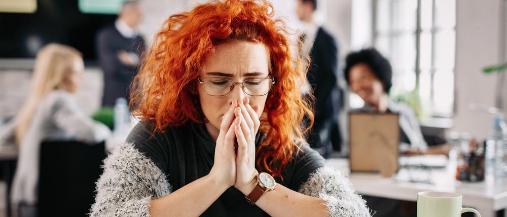 Dissatisfied businesswoman feeling sad and sitting at her office desk with eyes closed. There are people in the background.